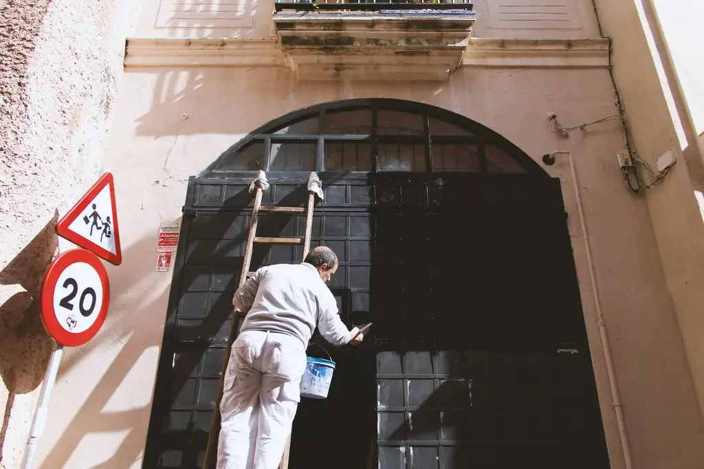 Peintre en tenue blanche sur une échelle, repeignant une grande porte cintrée sur une façade claire