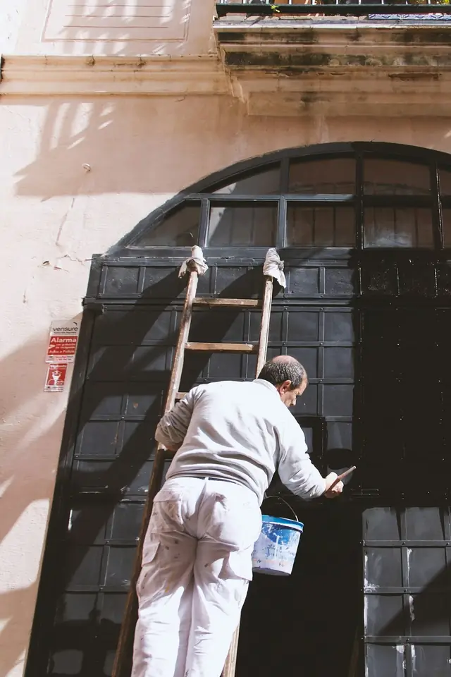 Peintre en tenue blanche sur une échelle, repeignant une grande porte cintrée sur une façade claire